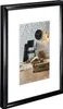 A black picture frame with a white matting, showcasing a framed image of a room interior with a chair, clock, and suitcase. Anteprima dell’immagine prodotto