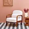 A cream-colored armchair with wooden legs and armrests sits on a black and white checkered floor in a room with pink walls. A small side table with a vase of dried flowers and a small bust is next to the chair. Produktbild-Vorschau
