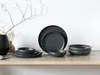 A set of dark gray stoneware plates and bowls arranged on a wooden shelf, with a decorative branch and vase in the background. Náhľad obrázku produktu