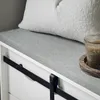 A close-up view of a white storage bench with a gray upholstered top and a black sliding barn door track. A white textured pillow rests on the bench, and a lit candle in a glass jar sits on a wooden coaster to the right. Anteprima dell’immagine prodotto