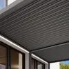 A close-up view of a gray aluminum pergola roof with a slatted design, attached to a white building with large windows. Voir les images du produit