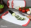 A set of three white cutting boards with red trim, one of which is in the foreground with a zucchini and knife on it. Other vegetables like tomatoes, onions, and lettuce are in the background. Anteprima dell’immagine prodotto