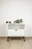 A white nightstand with a drawer and an open shelf, decorated with a few items on top, stands against a white wall. Podgląd zdjęcia produktu