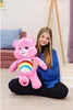 A young girl is sitting on the floor, holding a large pink plush teddy bear with a rainbow on its chest. Produktbild-Vorschau