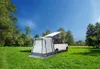 A white camper van with a pop-up roof and an attached awning is parked on a grassy field under a clear blue sky. Trees and distant mountains form the background. Produktbild-Vorschau