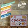 A close-up of a tin box with cookies depicted on it, placed on a wooden surface against a brick wall background. Text overlays describe product features. Voir les images du produit