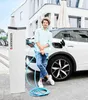 A man is standing next to a white electric car and a charging station, holding a charging cable. Náhľad obrázku produktu