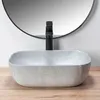 A close-up shot of a grey oval countertop sink with a black faucet, set against a white wall and a round mirror. Anteprima dell’immagine prodotto