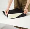 A person's hands are shown adjusting a black cushion with a white foam insert on a white table. Produktbild-Vorschau