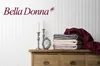 A stack of folded bedding in various colors sits on a wooden dresser, next to two silver candlesticks and a small ornate picture frame. The brand name 'Bella Donna' is displayed prominently on the wall above. Produktbild-Vorschau