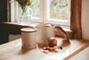 Two cream-colored storage containers with wooden lids and accents are on a wooden countertop next to a window. An onion and its peels are in the foreground. Produktbild-Vorschau