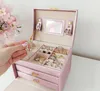 A hand is placing a silver necklace into an open pink jewelry box, which is filled with various rings. A bouquet of pink flowers is in the background. Náhled obrázku produktu