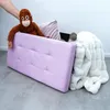 A hand is placing a stuffed orangutan on top of a purple storage bench, which is next to a white fluffy blanket and a small gray stuffed hippo. Náhled obrázku produktu