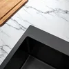 A close-up view of a black sink on a marble countertop, with a wooden cutting board in the upper left corner. Produktbild-Vorschau