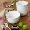 A stack of white bowls with a spoon, limes, and a napkin on a wooden table. Produktbild-Vorschau