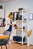 A woman is standing in a kitchen or office space next to a white shelving unit. Produktbild-Vorschau