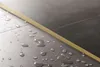 A close-up view of a gold-colored profile strip separating two sections of gray tile, with water droplets scattered on the tiles. Produktbild-Vorschau