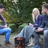 Three people are sitting around a fire pit, holding mugs and talking. Produktbild-Vorschau