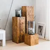 A collection of wooden pedestals of varying heights, some topped with decorative items like glass vases with stones and a candle holder with a candle. A framed picture leans against the wall in the background. Produktbild-Vorschau