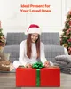 A woman in a Santa hat is sitting on the floor in front of a gray sofa, opening a large red gift box with a green ribbon. Christmas trees are visible in the background. Produktbild-Vorschau