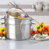 A set of stainless steel pots with a steamer basket and a glass lid, arranged on a kitchen counter with vegetables and herbs. Produktbild-Vorschau