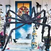 A Halloween decoration scene featuring a large, furry spider, stacked skulls, and floral arrangements in front of a blue door. Produktbild-Vorschau