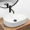 A white oval countertop sink with a black faucet is shown on a wooden countertop, with a reflection in a round mirror behind it. Anteprima dell’immagine prodotto