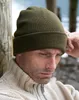 A man wearing a green beanie and a cream-colored sweater, leaning against a wooden post with a body of water in the background. Anteprima dell’immagine prodotto