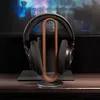 A pair of black headphones rests on a wooden and metal headphone stand, placed on a wooden desk in front of a monitor displaying a colorful, abstract image. Produktbild-Vorschau