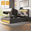 A modern bedroom with a grey upholstered bed frame, partially open to reveal storage space underneath. The bed has LED lighting along the base and headboard. A tablet is placed on the headboard, and a large framed artwork hangs on the wall above. Náhľad obrázku produktu