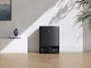 A black robot vacuum cleaner with its docking station is placed on a wooden floor against a white wall. Sunlight casts shadows on the wall. Produktbild-Vorschau