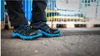 A person wearing black pants and black and blue safety shoes standing on a concrete surface with a blurred background of industrial equipment. Náhľad obrázku produktu