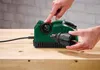 A person's hands are shown operating a green drill sharpener on a wooden workbench. Produktbild-Vorschau