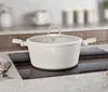A cream-colored pot with a gold handle sits on a black induction cooktop, with a blurred kitchen background. Produktbild-Vorschau