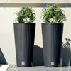 Two tall, dark grey planters with green plants inside, placed on a wooden deck against a light-colored wall. Produktbild-Vorschau