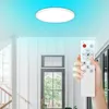 A hand holding a remote control is shown in the foreground, with a modern LED ceiling light illuminating the ceiling with a gradient of blue light in the background. A doorway and staircase are also visible. Produktbild-Vorschau