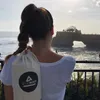 A woman stands with her back to the camera, looking out at a scenic ocean view with a temple on a rock formation. She is carrying a yoga mat bag over her shoulder. Produktbild-Vorschau
