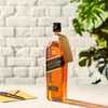 A bottle of Johnnie Walker Black Label whisky sits on a table with an envelope and pen. Produktbild-Vorschau
