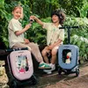 Two children are sitting on a bench in a garden, playing with toy tea sets and their ride-on suitcases. Produktbild-Vorschau