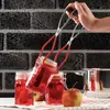A hand holding a jar lifter with a jar of red liquid, with other jars and an apple in the foreground. Anteprima dell’immagine prodotto