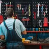 A person in overalls stands in front of a pegboard wall filled with tools in a workshop. Produktbild-Vorschau