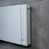 A white radiator panel is mounted on a gray tiled wall. Produktbild-Vorschau