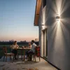 A family is sitting at a table on a patio at dusk, with outdoor wall lights illuminating the scene. Produktbild-Vorschau