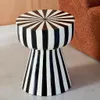 A black and white striped stool is placed on a light gray floor next to a brown textured object. Produktbild-Vorschau