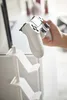 A hand holding a white game controller near a white stand with multiple shelves. Produktbild-Vorschau