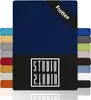 A product image showcasing a navy blue fitted sheet with a black band at the top corner that says 'Frottee'. To the left and right of the main sheet are stacks of fitted sheets in various colors. Produktbild-Vorschau