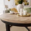 A close-up of a round coffee table with a wood-grain top and black metal legs, set in a living room with a vase of yellow flowers, a cup of coffee, and candles. Produktbild-Vorschau