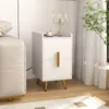 A white nightstand with gold legs and handle is placed next to a sofa, with a lamp and a cup on top. A curtain and a plant are visible in the background. Produktbild-Vorschau