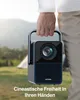 A person is holding a portable projector outdoors, with tents and trees in the background. Produktbild-Vorschau