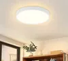 A white, circular LED ceiling light is mounted on a white ceiling. Below the light, a wooden shelf holds a potted plant and some books. Produktbild-Vorschau